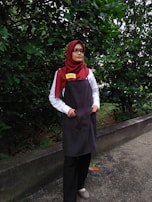 A person is standing outdoors in front of dense green foliage. They are wearing a maroon headscarf, glasses, a white shirt, a dark apron, and black pants. The person has a name tag that reads 'DINIE'. The background consists of a lush green hedge and a concrete path.
