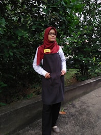 A person is standing outdoors in front of dense green foliage. They are wearing a maroon headscarf, glasses, a white shirt, a dark apron, and black pants. The person has a name tag that reads 'DINIE'. The background consists of a lush green hedge and a concrete path.