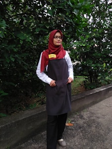 A person is standing outdoors in front of dense green foliage. They are wearing a maroon headscarf, glasses, a white shirt, a dark apron, and black pants. The person has a name tag that reads 'DINIE'. The background consists of a lush green hedge and a concrete path.