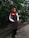 A person is standing outdoors in front of dense green foliage. They are wearing a maroon headscarf, glasses, a white shirt, a dark apron, and black pants. The person has a name tag that reads 'DINIE'. The background consists of a lush green hedge and a concrete path.