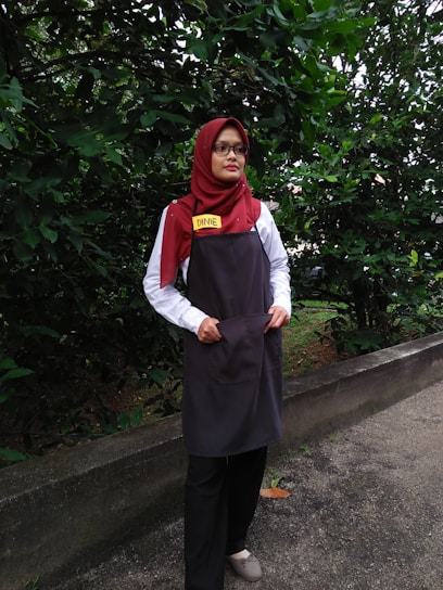 A person is standing outdoors in front of dense green foliage. They are wearing a maroon headscarf, glasses, a white shirt, a dark apron, and black pants. The person has a name tag that reads 'DINIE'. The background consists of a lush green hedge and a concrete path.