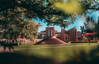 A well-maintained green lawn surrounds a central architectural structure with steps and columns. People are walking and sitting around, enjoying the outdoor space. Overhead, trees provide shade, and there is a clear blue sky in the background, adding a serene atmosphere to the scene.