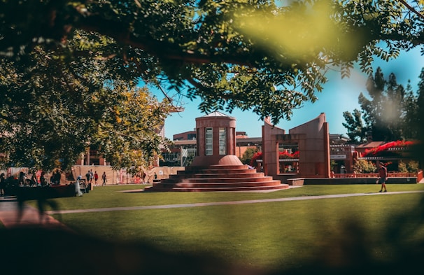 A well-maintained green lawn surrounds a central architectural structure with steps and columns. People are walking and sitting around, enjoying the outdoor space. Overhead, trees provide shade, and there is a clear blue sky in the background, adding a serene atmosphere to the scene.
