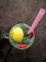 A refreshing beverage is presented in a glass mug on a wooden surface. The drink contains a whole lemon, small seeds dispersed throughout, and pieces of red peppers. A pink straw is placed in the mug, suggesting it is ready to be consumed.