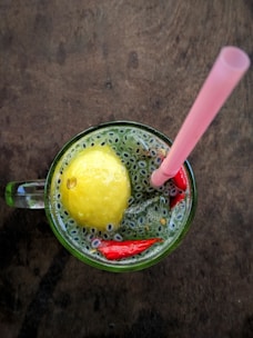 A refreshing beverage is presented in a glass mug on a wooden surface. The drink contains a whole lemon, small seeds dispersed throughout, and pieces of red peppers. A pink straw is placed in the mug, suggesting it is ready to be consumed.