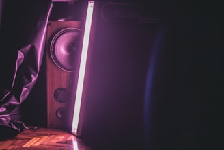 A wooden speaker with visible audio drivers is illuminated by a vertical, bright neon pink light. The scene is set against a dark background, with part of the image displaying a crumpled, dark sheet or fabric to the left. The lighting casts a soft glow and creates contrasting shadows.