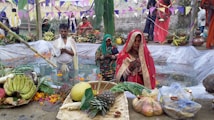 People wearing traditional clothing are participating in a religious ceremony near a small water body. There are offerings including fruits, flowers, and fire arranged around the water. Banners and flags are hung above the area, adding a festive atmosphere.
