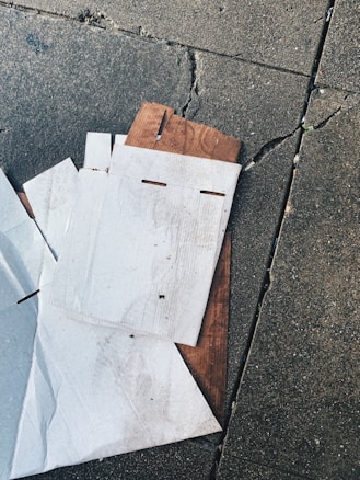 Several pieces of flattened cardboard are lying on a cracked concrete surface. The cardboard appears used and is slightly dirty. It is layered on the ground with some wooden material under it.