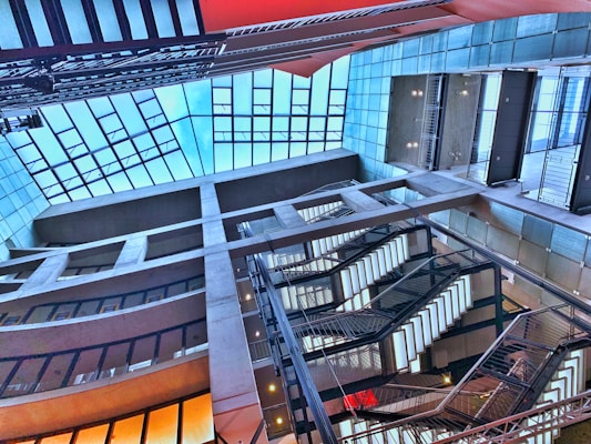 A modern architectural interior featuring a multi-level atrium with large glass windows and several staircases. The design incorporates a grid-like structure with concrete beams and metal railings, complemented by vibrant lighting.
