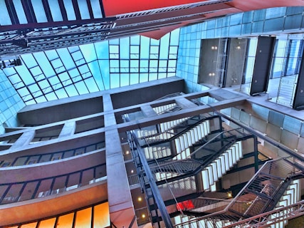 A modern architectural interior featuring a multi-level atrium with large glass windows and several staircases. The design incorporates a grid-like structure with concrete beams and metal railings, complemented by vibrant lighting.