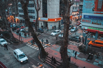 A busy urban street scene with numerous pedestrians walking along sidewalks and crossing streets. Cars and a taxi navigate through the intersection, bordered by large trees and surrounding buildings. Businesses with colorful signs are visible, including a bank and a language school.