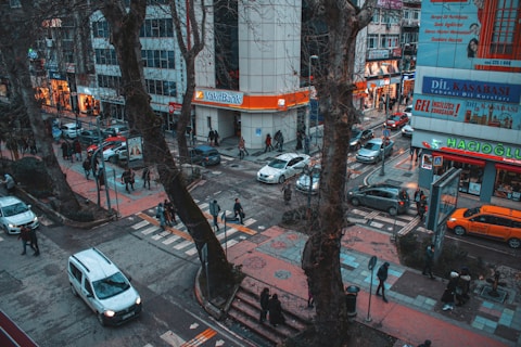A busy urban street scene with numerous pedestrians walking along sidewalks and crossing streets. Cars and a taxi navigate through the intersection, bordered by large trees and surrounding buildings. Businesses with colorful signs are visible, including a bank and a language school.