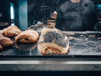 A selection of freshly baked pastries, including croissants and a poppy-seed roll, arranged on a metal tray. The background shows a person partially obscured and blurred, likely behind a glass display case in a bakery setting.