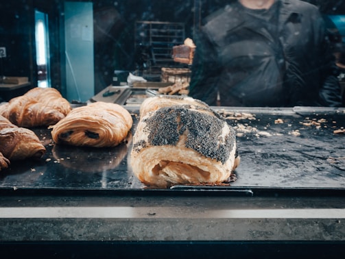 A selection of freshly baked pastries, including croissants and a poppy-seed roll, arranged on a metal tray. The background shows a person partially obscured and blurred, likely behind a glass display case in a bakery setting.