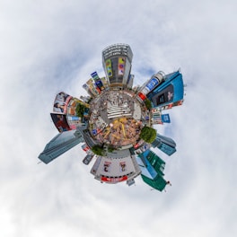 A creative 'tiny planet' view of an urban cityscape featuring a bustling intersection surrounded by tall buildings with colorful advertisements. The scene captures numerous people crossing the street, with vibrant billboards on various skyscrapers adding to the city's energetic atmosphere.