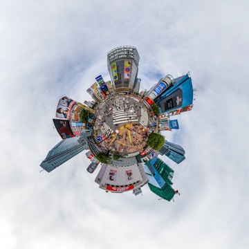 A creative 'tiny planet' view of an urban cityscape featuring a bustling intersection surrounded by tall buildings with colorful advertisements. The scene captures numerous people crossing the street, with vibrant billboards on various skyscrapers adding to the city's energetic atmosphere.
