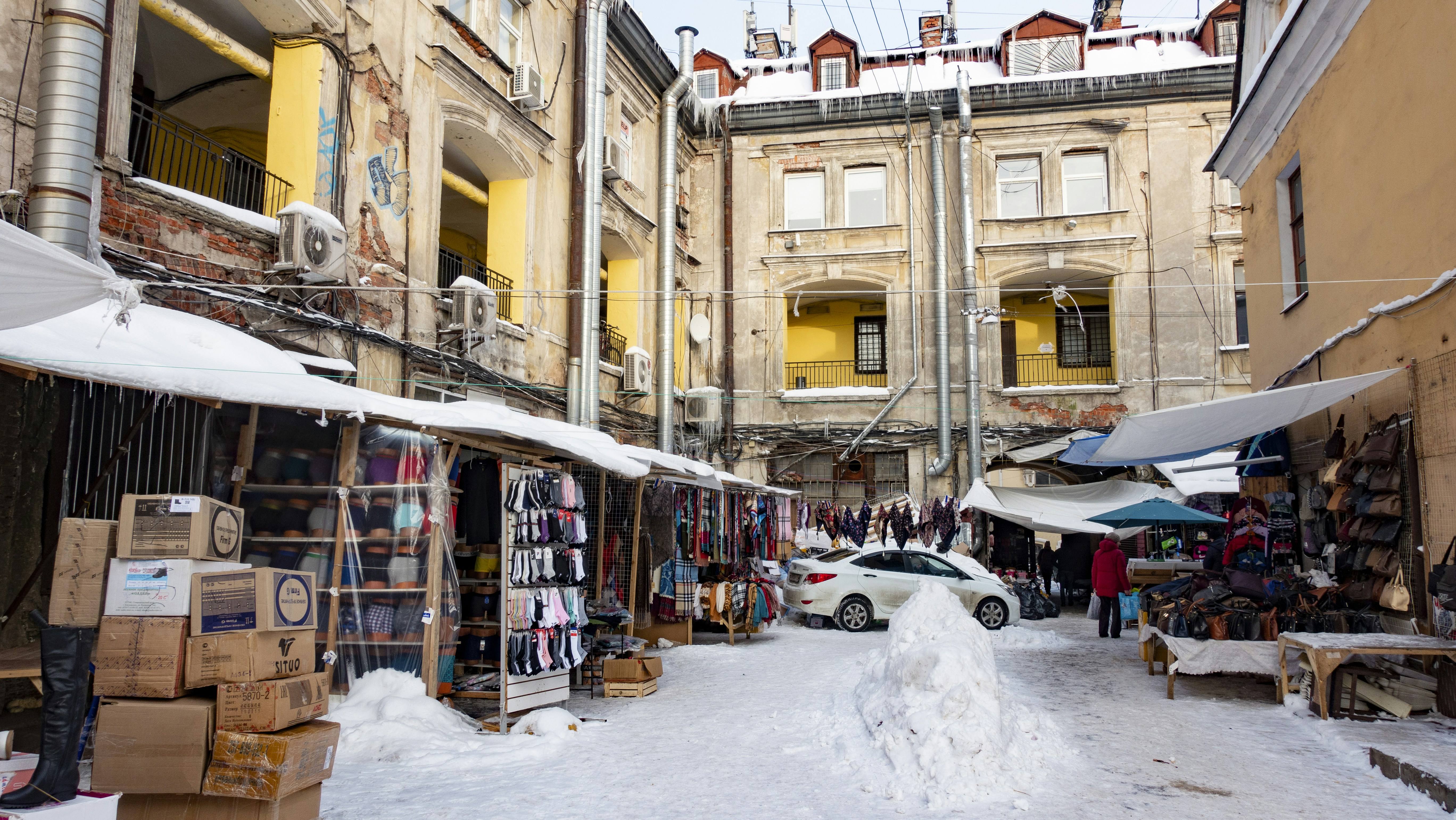 A marketplace set in an urban courtyard surrounded by old buildings with visible signs of wear. Stalls line the sides, displaying various goods such as clothing and accessories. A covering of snow is visible on the ground and stalls. A parked car is near the center, and several people are present, suggesting a bustling environment.