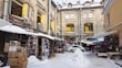 A marketplace set in an urban courtyard surrounded by old buildings with visible signs of wear. Stalls line the sides, displaying various goods such as clothing and accessories. A covering of snow is visible on the ground and stalls. A parked car is near the center, and several people are present, suggesting a bustling environment.