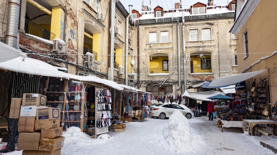 A marketplace set in an urban courtyard surrounded by old buildings with visible signs of wear. Stalls line the sides, displaying various goods such as clothing and accessories. A covering of snow is visible on the ground and stalls. A parked car is near the center, and several people are present, suggesting a bustling environment.