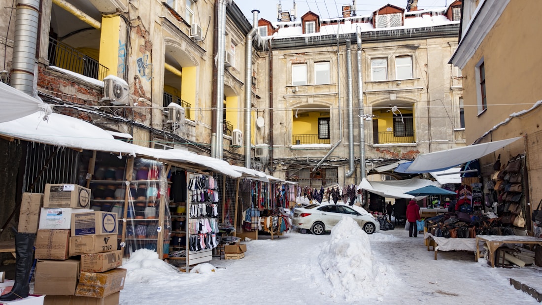 A marketplace set in an urban courtyard surrounded by old buildings with visible signs of wear. Stalls line the sides, displaying various goods such as clothing and accessories. A covering of snow is visible on the ground and stalls. A parked car is near the center, and several people are present, suggesting a bustling environment.