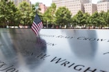 A small American flag stands on a memorial inscribed with names. In the background, there are trees and buildings, indicating an outdoor setting in an urban environment.
