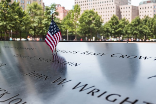 A small American flag stands on a memorial inscribed with names. In the background, there are trees and buildings, indicating an outdoor setting in an urban environment.