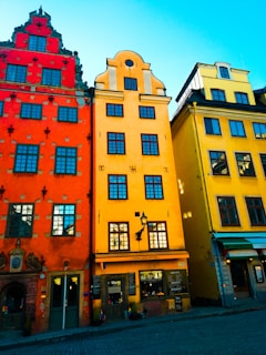 Vibrant, colorful buildings line a quaint cobblestone street. The architecture features intricate designs and distinct shapes, with a bright blue sky overhead.