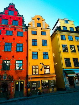 Vibrant, colorful buildings line a quaint cobblestone street. The architecture features intricate designs and distinct shapes, with a bright blue sky overhead.