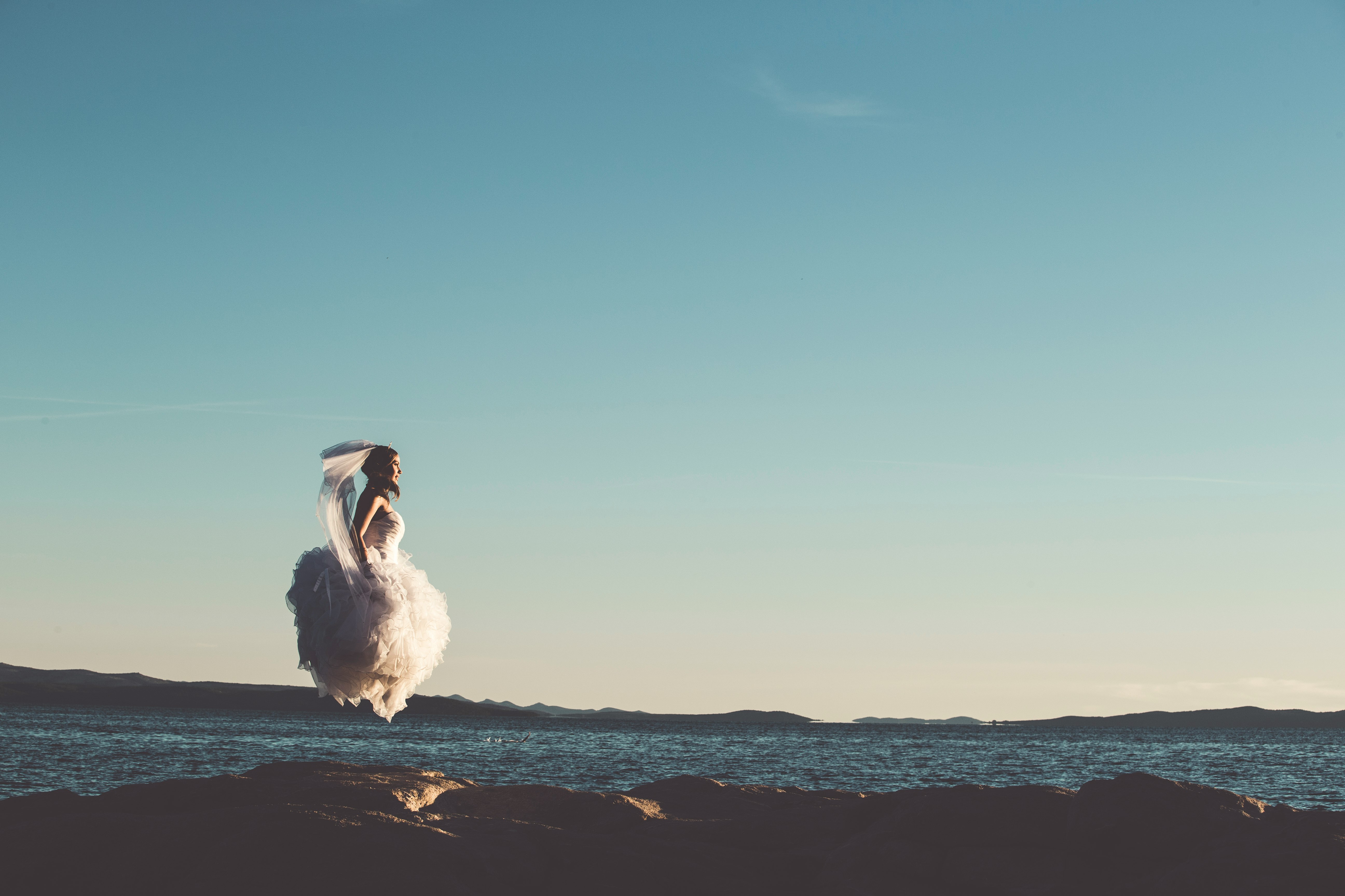woman in white bridal gown jump in mid air next to body of water