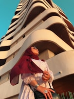 A person wearing a red hijab and sunglasses stands in front of a modern high-rise building with wave-like architectural features. The person is dressed in a patterned dress and accessorized with rings and bracelets. The sky is clear and blue, providing a striking contrast to the earth-toned structure.
