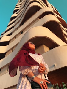 A person wearing a red hijab and sunglasses stands in front of a modern high-rise building with wave-like architectural features. The person is dressed in a patterned dress and accessorized with rings and bracelets. The sky is clear and blue, providing a striking contrast to the earth-toned structure.