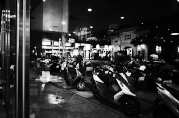 A row of parked scooters is visible alongside reflective glass windows that mirror the bustling street scene. The background features illuminated signage and an entrance to a building, hinting at an urban environment, possibly a shopping area or market.