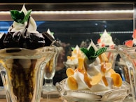 A display of two intricately prepared dessert glasses featuring soft serve ice cream. The dessert on the left includes dark chocolate pieces and is topped with mint leaves, while the dessert on the right is garnished with caramel sauce and almond slices, also adorned with mint leaves.