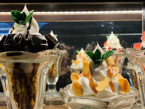 A display of two intricately prepared dessert glasses featuring soft serve ice cream. The dessert on the left includes dark chocolate pieces and is topped with mint leaves, while the dessert on the right is garnished with caramel sauce and almond slices, also adorned with mint leaves.