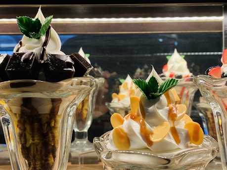 A display of two intricately prepared dessert glasses featuring soft serve ice cream. The dessert on the left includes dark chocolate pieces and is topped with mint leaves, while the dessert on the right is garnished with caramel sauce and almond slices, also adorned with mint leaves.