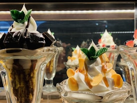A display of two intricately prepared dessert glasses featuring soft serve ice cream. The dessert on the left includes dark chocolate pieces and is topped with mint leaves, while the dessert on the right is garnished with caramel sauce and almond slices, also adorned with mint leaves.