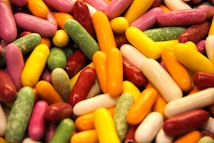 A vibrant assortment of elongated, cylindrical candies in various colors, including pink, yellow, green, white, and orange, scattered together creating a playful and colorful texture.