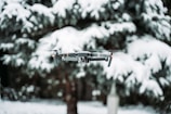 A drone hovers in front of snow-covered trees, which create a blurred wintery background. The device is mid-flight, showcasing propellers and a camera mounted at the front.