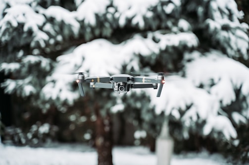 A drone hovers in front of snow-covered trees, which create a blurred wintery background. The device is mid-flight, showcasing propellers and a camera mounted at the front.