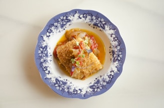 A beautifully plated dish featuring a piece of breaded and fried food, possibly fish or meat, garnished with microgreens and placed in a decorative blue and white floral patterned bowl. The dish is accompanied by vibrant vegetables and a light sauce.