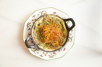 A round, black-handled dish is filled with a creamy baked pasta topped with crispy fried shoe string fries and green herbs. The dish sits on a floral patterned plate with a white background.