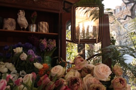 A rustic setting filled with a variety of flowers, including roses and tulips, placed in a wooden structure with shelves. A decorative ceramic bird is perched on the top shelf alongside patterned vases. Sunlight filters through a window, casting a warm glow over the scene and highlighting the lush greenery and vibrant flowers.