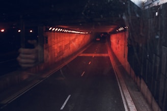 A dimly lit tunnel with orange lighting along the walls, displaying a sense of depth as it extends into darkness. There is a reflective surface showing a hand holding a phone. The tunnel's interior is lined with lights creating an industrial atmosphere, while a lone vehicle is visible in the distance.