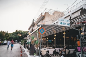 An outdoor café with a sign reading 'Greek Art,' displaying several empty chairs and tables under a covered area. The structure is attached to a multi-story building with balconies and metal railings. A few people walk along the adjacent street lined with orange traffic cones, while tall green trees are visible in the background. The atmosphere suggests an early evening setting.