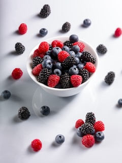 A white bowl filled with a mix of fresh blackberries, blueberries, and raspberries sits on a white surface. More berries are scattered around the bowl in a casual arrangement.
