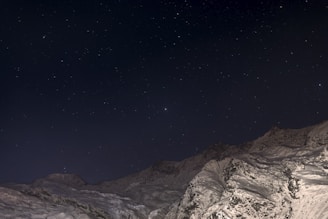 A panoramic view of towering mountains with snow melting into a pristine river, framed by a deep midnight black sky.