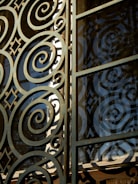 Intricate metalwork featuring spirals and geometric patterns integrated into a gate or window. The structure is crafted with ornate, swirling designs, casting shadows and reflections against a glass panel.