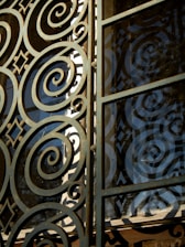 Intricate metalwork featuring spirals and geometric patterns integrated into a gate or window. The structure is crafted with ornate, swirling designs, casting shadows and reflections against a glass panel.