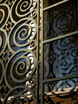 Intricate metalwork featuring spirals and geometric patterns integrated into a gate or window. The structure is crafted with ornate, swirling designs, casting shadows and reflections against a glass panel.
