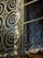 Intricate metalwork featuring spirals and geometric patterns integrated into a gate or window. The structure is crafted with ornate, swirling designs, casting shadows and reflections against a glass panel.